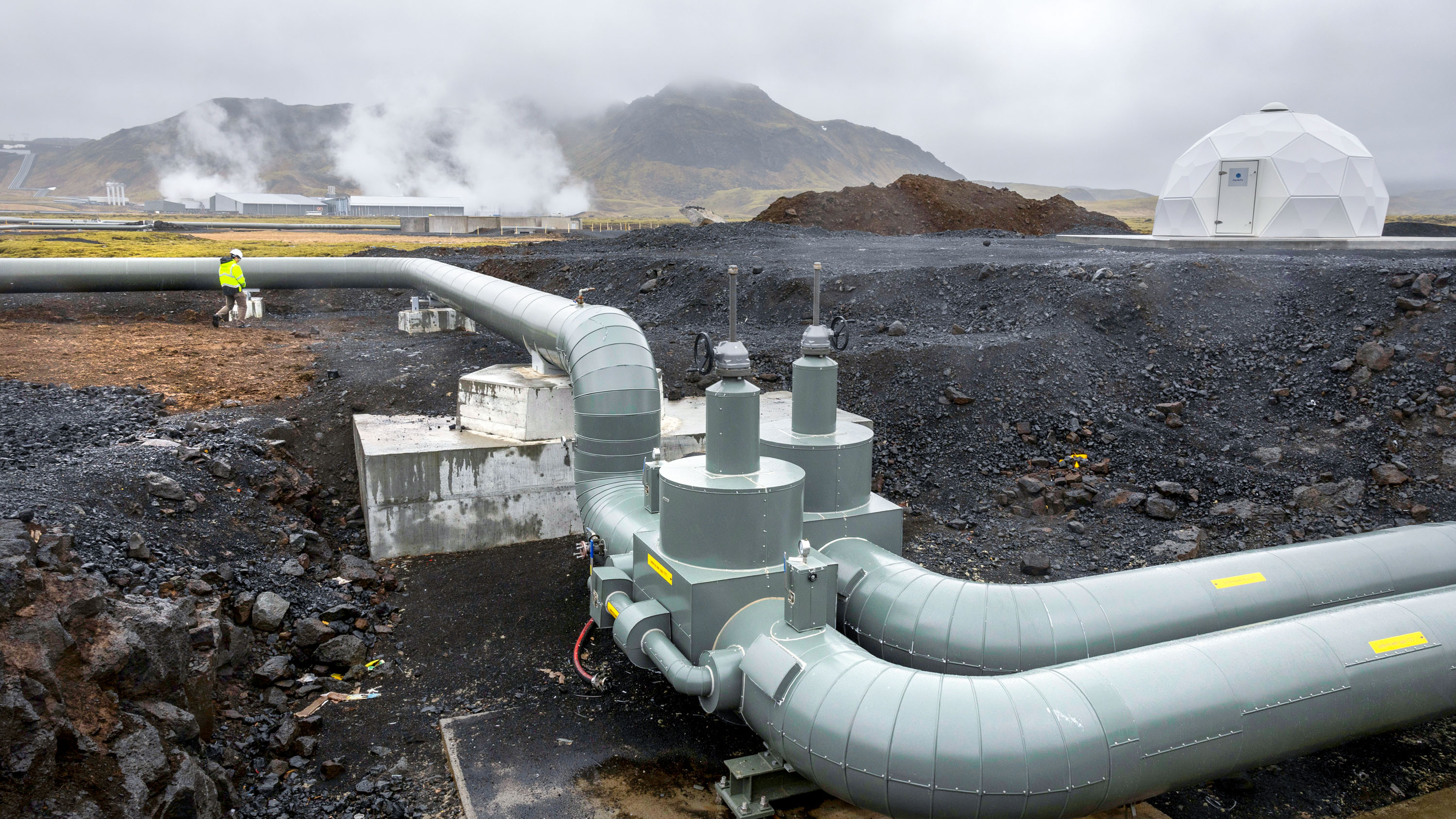 large pipes run along the ground with the buildings of the Climeworks' Mammoth plant in the distance