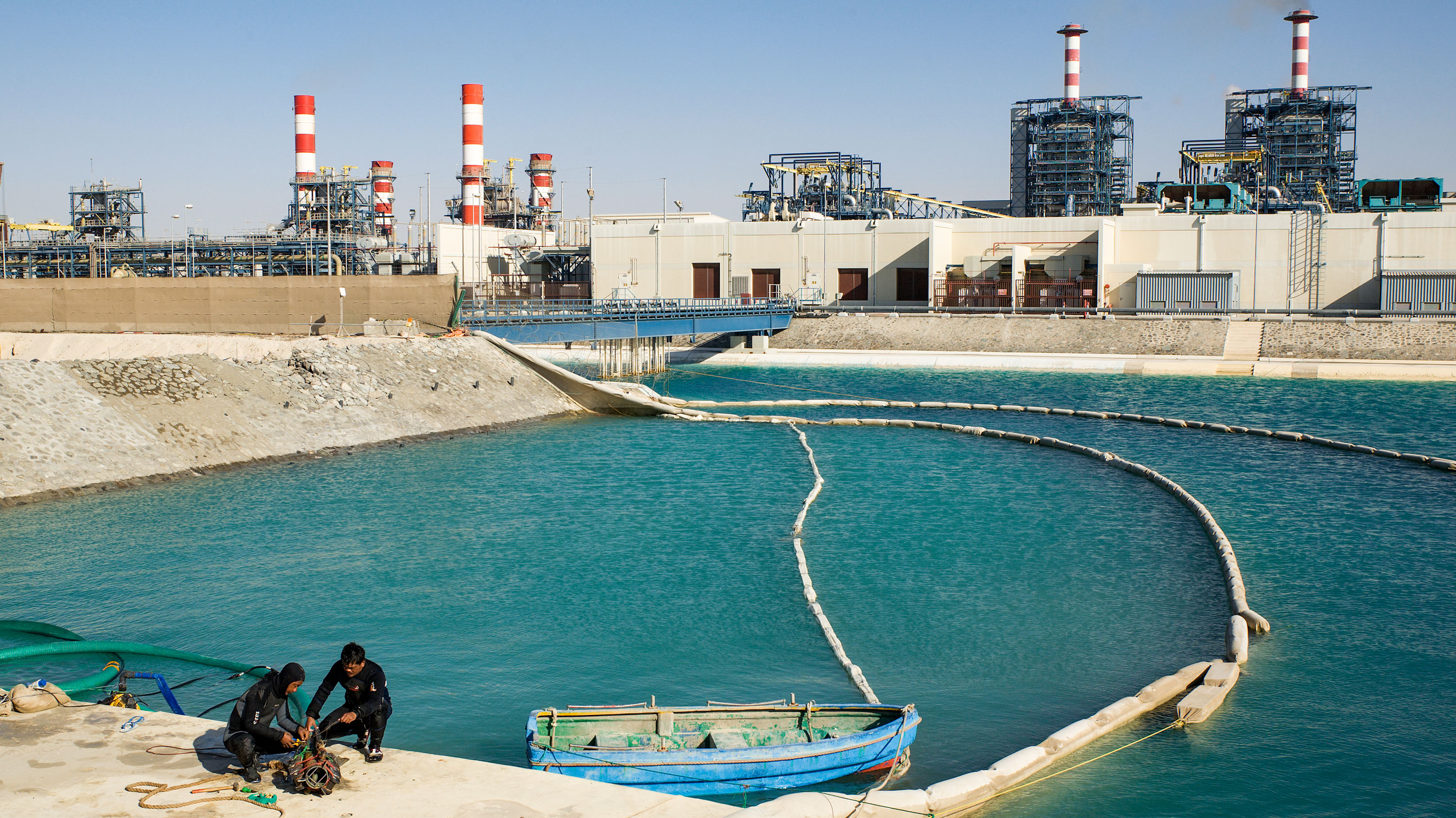 In the foreground, workers in scuba gear crouch next to a rowboat near the sea water intake pool of the desalination plant in the background.