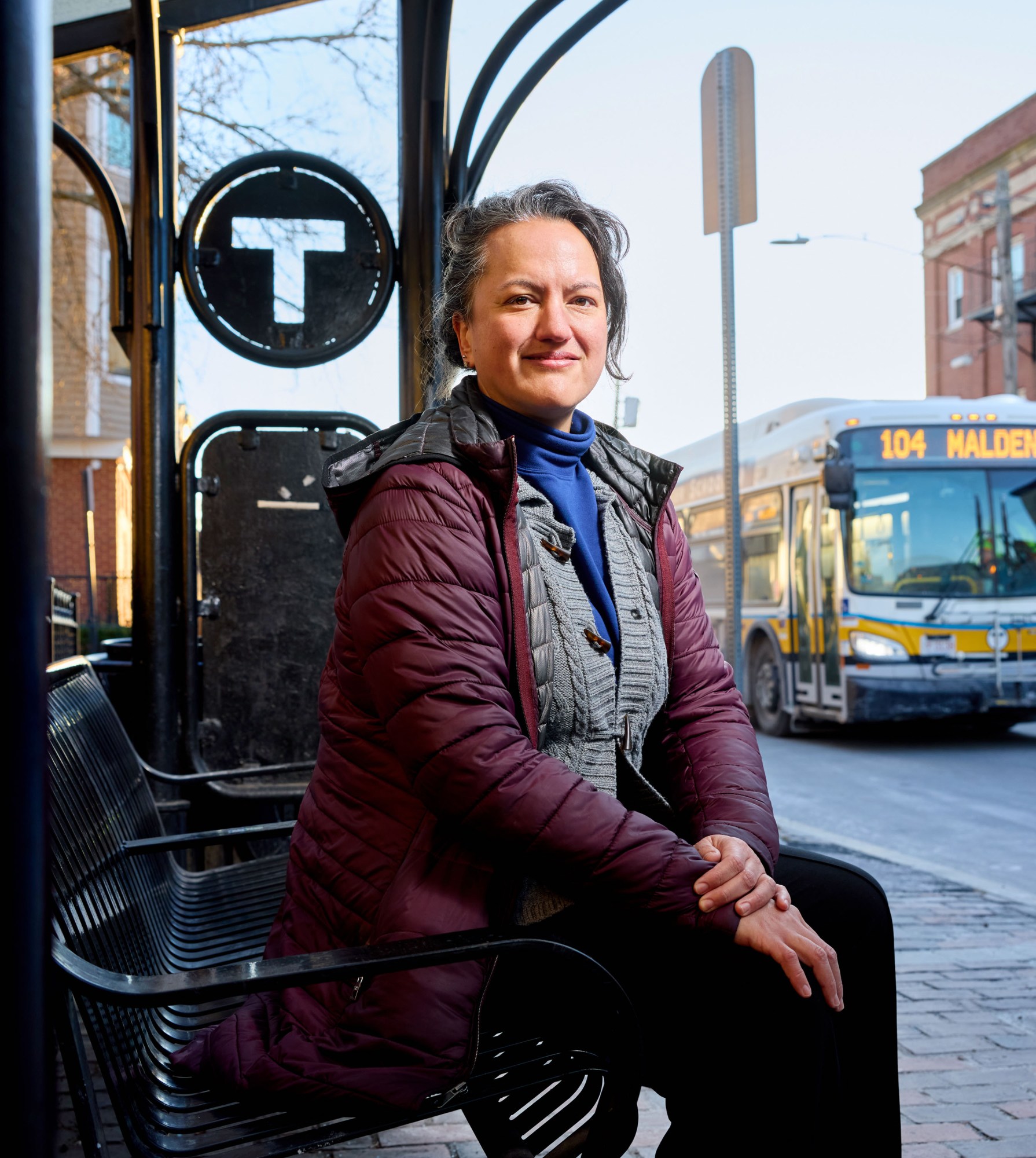 Melissa Dullea sitting at a bus stop near a 104 bus to Malden